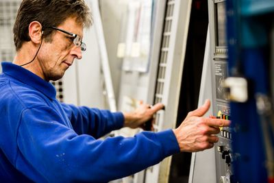A side-profile photograph shows a middle-aged man wearing glasses and a blue sweatshirt, pressing a red button on the control panel of a large industrial machine.