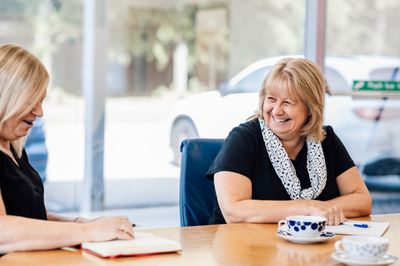 Two professional female Heald employees smiling and laughing during a meeting at a conference table with tea and notebooks.