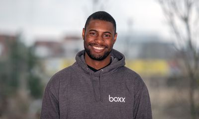 A smiling man looking directly at the camera, wearing a dark grey hoodie with a white 'boxx' logo on the left chest, set against a softly blurred outdoor background.
