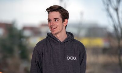 A smiling man looking off-camera to the left, wearing a dark grey hoodie with a white 'boxx' logo on the left chest, set against a softly blurred outdoor background.