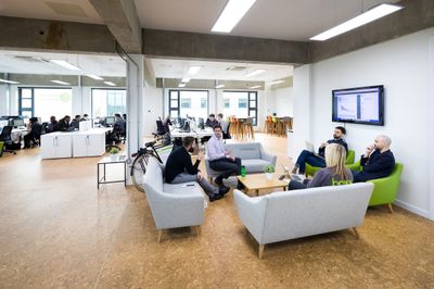 A wide view of a spacious, open-plan office with cork flooring. Several employees are gathered in a casual meeting area featuring grey sofas and green armchairs, while others work at rows of white desks in the background.