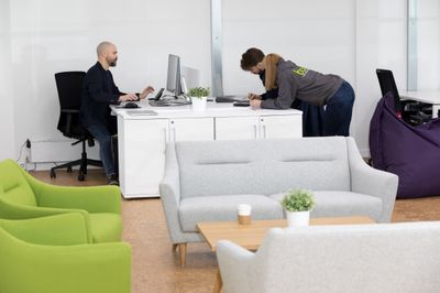 Three colleagues collaborating at a white desk in a modern office space. One person is seated at a computer monitor, while two others lean over the desk to review materials. One person is wearing a grey 'boxx' branded hoodie.