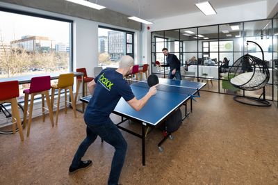 Two men play ping pong in a modern, brightly lit office with a cork floor. The man in the foreground, wearing a dark blue t-shirt with the lime green 'boxx' logo, reaches to hit the ball, while the man on the other side smiles. Large windows with a view of other buildings line one wall, and a glass-partitioned meeting room with other employees, a bar with colorful stools, and a hanging swing chair are in the background.