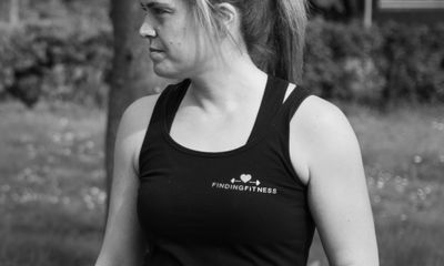 A black and white photograph of a woman looking off to the side, wearing a black tank top. The top features the 'Finding Fitness' logo on the left chest, which includes a barbell with a heart in the center.