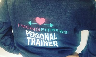 A close-up view of the back of a dark grey sweatshirt. It features a large "Finding Fitness" logo with a pink heart and teal barbell, positioned above large, bold white text that reads "PERSONAL TRAINER". The person wearing it has their hands resting on their hips.