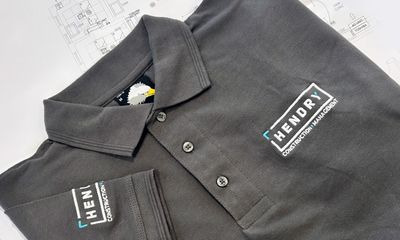 A charcoal grey polo shirt with hendry Logo embroidered in white and teal on the left chest and right sleeve. The shirt is laid flat on top of architectural blueprints showing a floor plan.