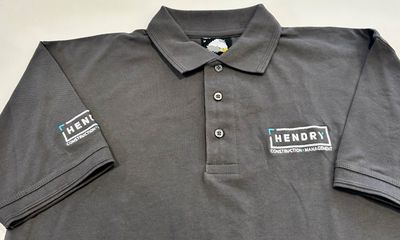 A front-facing, flat-lay view of a charcoal grey polo shirt. The shirt features three buttons and an embroidered company logo on the chest and sleeve.