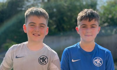 Two boys standing side-by-side outdoors, wearing MK United branded soccer training shirts—one in grey and one in blue.