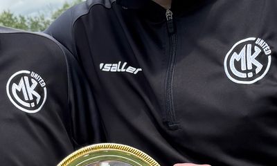 A close-up of a black quarter-zip sports jacket featuring the white embroidered MK UNITED circular logo on the chest and sleeve.