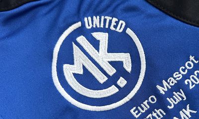 A macro shot of a blue soccer jersey showing a thick, white embroidered MK UNITED circular logo, with partial white text stitched below it.