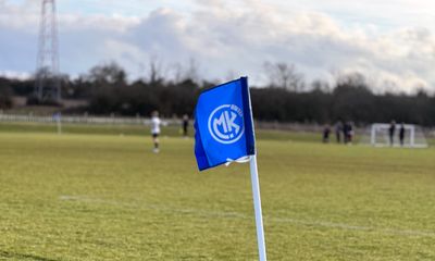 A blue soccer corner flag with a white MK UNITED logo blowing in the wind on a green pitch, with blurred players in the background.