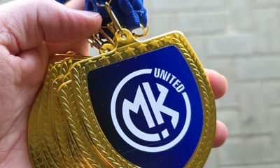 A hand holding a cluster of shiny gold medals on blue ribbons. Each medal has a blue center insert displaying the white MK UNITED logo.