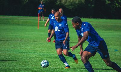 A daytime football match on a green field, featuring two male players in blue kits sprinting after the ball. The player on the left has tattoos on his leg. Other players and a coach are visible in the background, along with a line of trees.