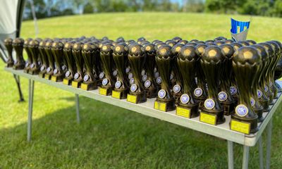MK United presentation day - A long table outdoors on a grass field is filled with dozens of identical dark soccer trophies, each featuring a soccer ball at the top and a blue circular MK United logo on the base. A single blue and silver cup-style trophy is visible at the far right end of the table.