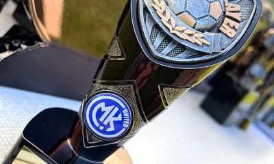 A close-up of a black, silver, and gold soccer trophy that has a small, circular MK UNITED logo emblem attached to its stem.