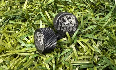 Two small, black metal thumbscrews with textured, knurled edges resting on artificial green grass. The flat top of each screw is laser-engraved with the 'Nero Angling' logo, which features a leaping fish.